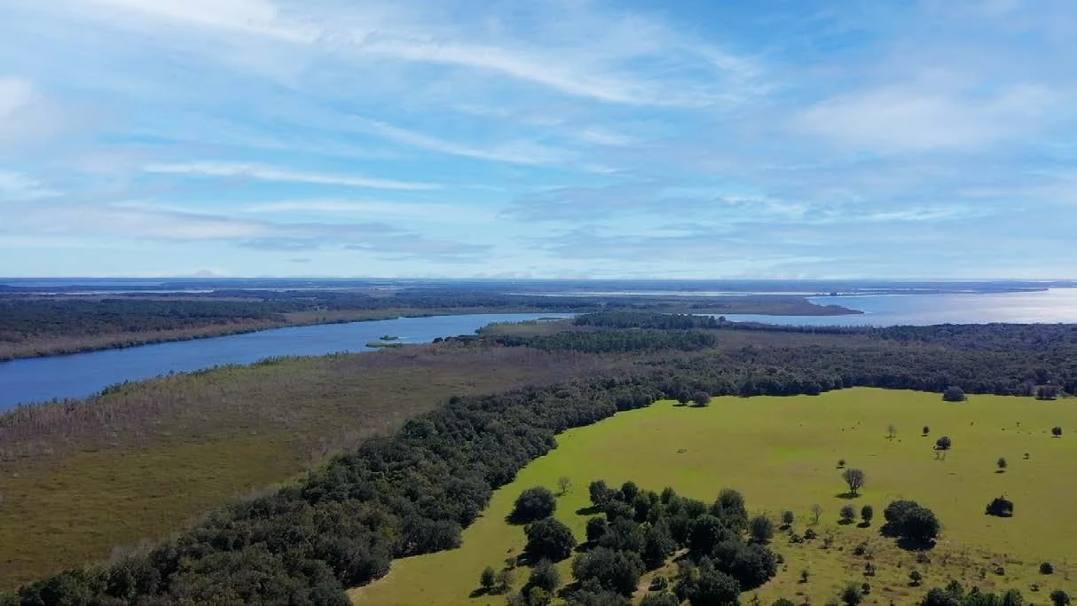 Aerial view across pasture and wooded shoreline toward surrounding waterways